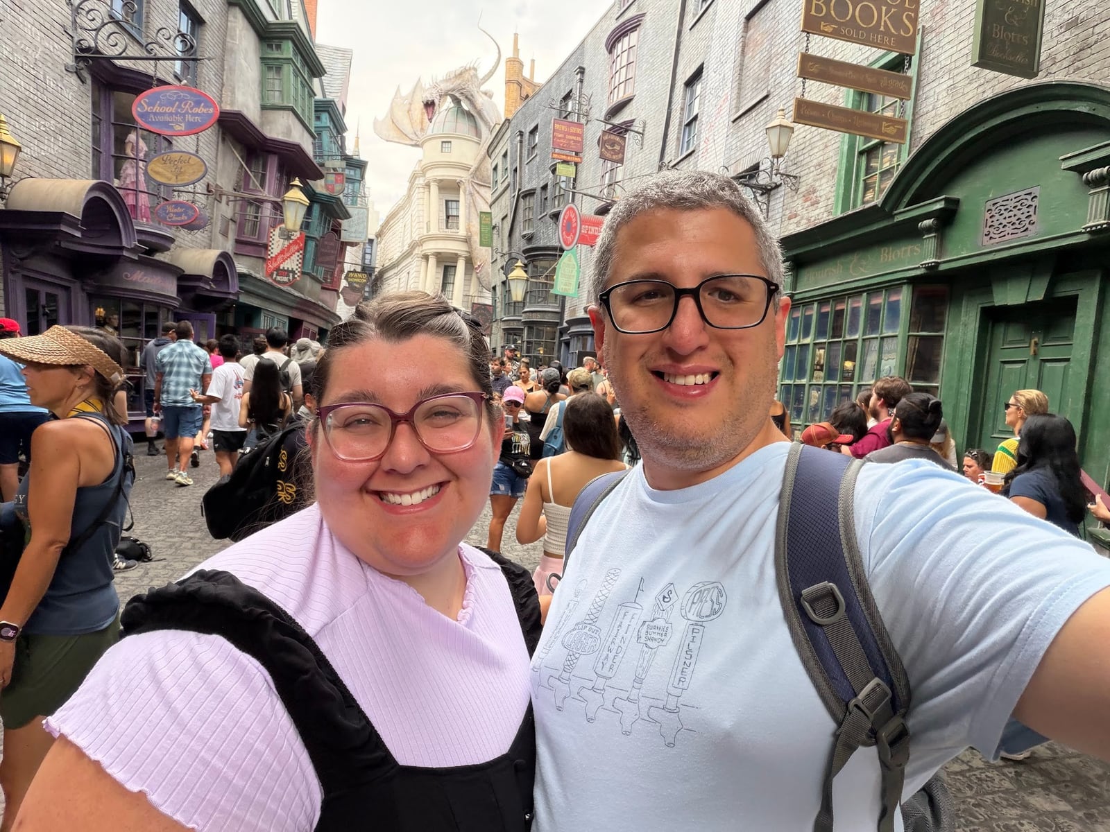 Two smiling travelers pose for a selfie in a crowded themed street area with colorful buildings and shops.